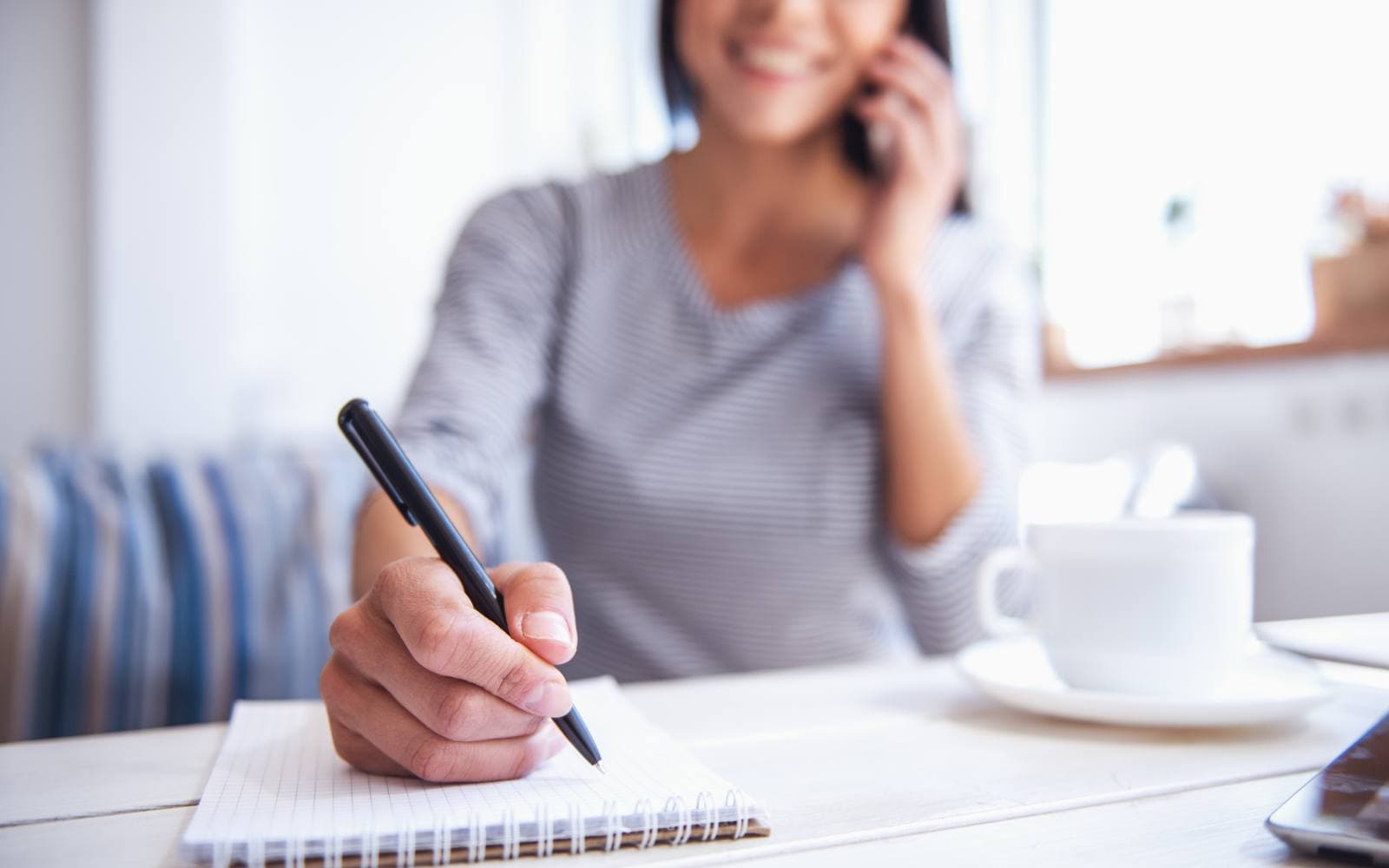 A smiling woman on a phone call, writing notes with a pen in a spiral notebook at a white table beside a cup of coffee