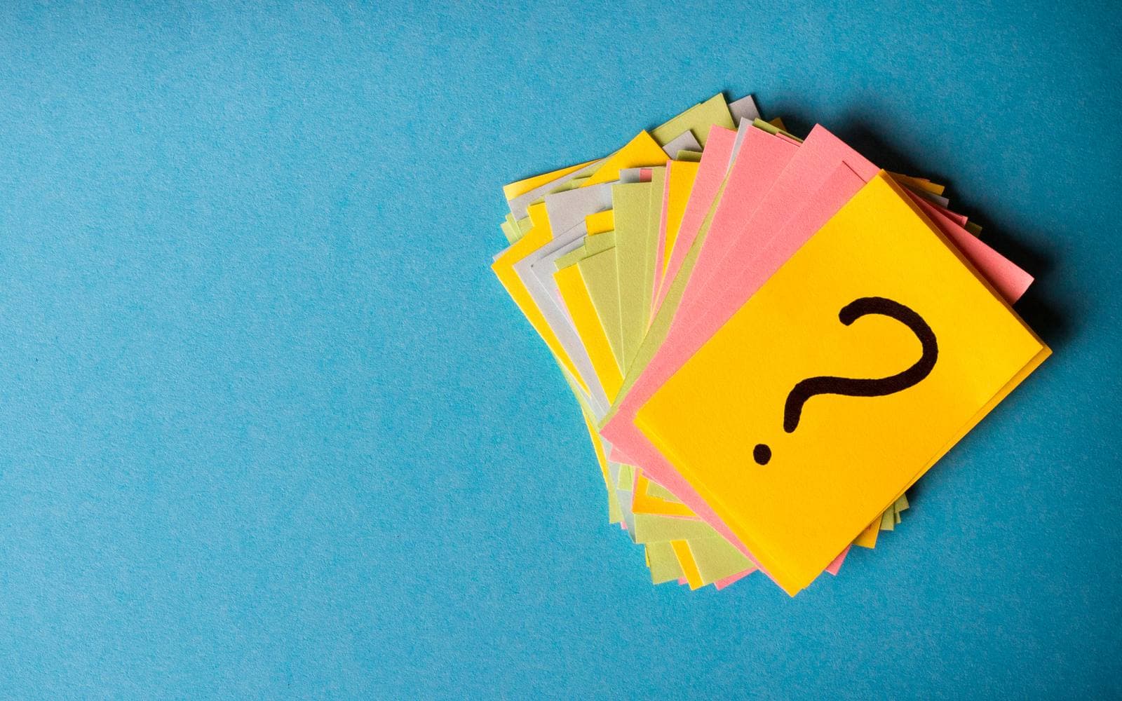 A fanned stack of colourful paper cards on a blue surface, the top card yellow and marked with a hand-drawn question mark
